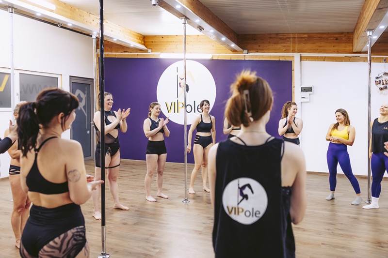 Zeigt eine Gruppe lachender Frauen im Kreis aufgestellt umringt von Poledance Stangen